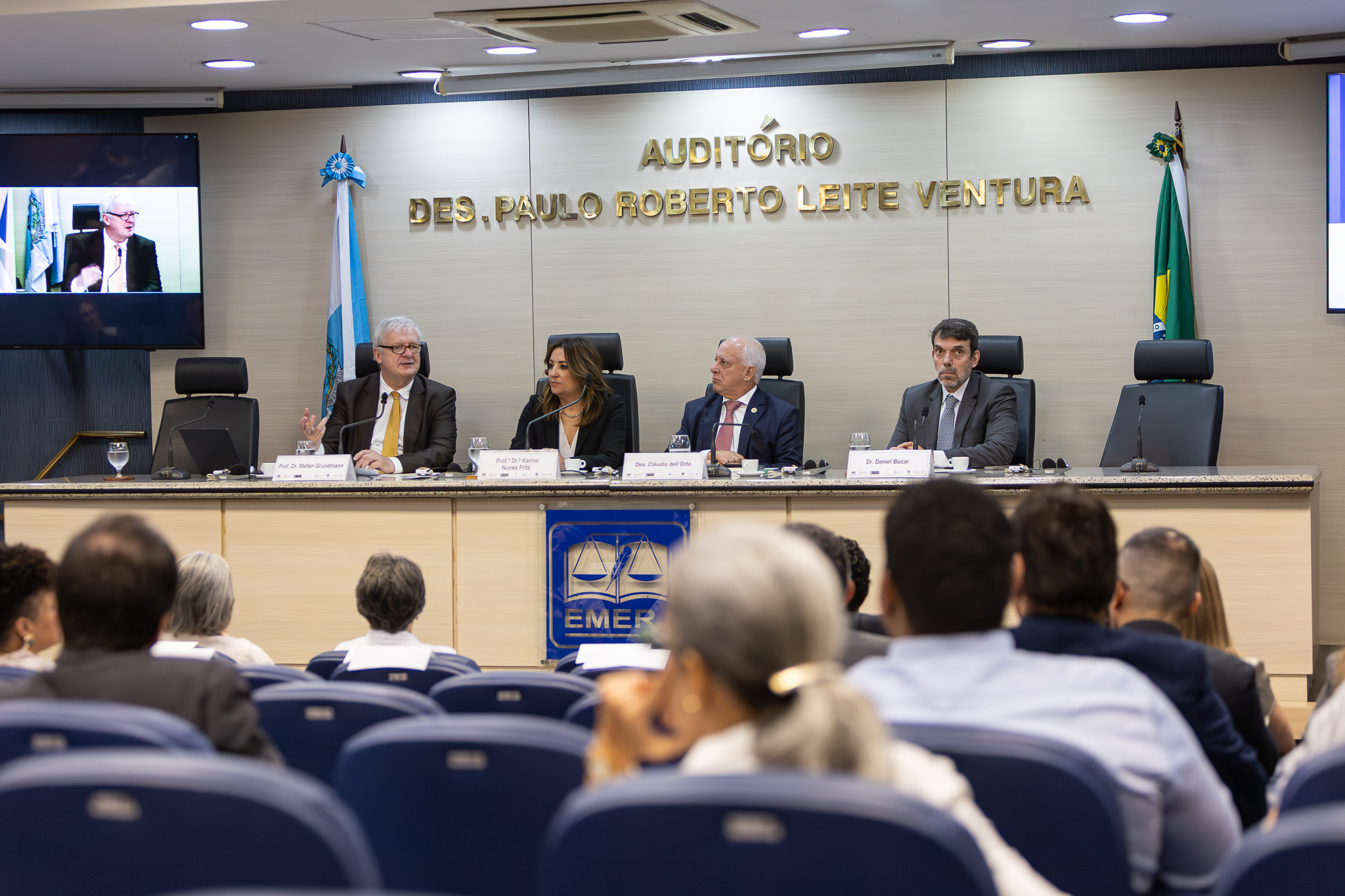 A imagem mostra um evento institucional realizado em um auditório, com uma mesa de autoridades posicionada à frente e público sentado na plateia. Ao fundo, na parede, está escrito em letras douradas: “AUDITÓRIO DES. PAULO ROBERTO LEITE VENTURA”. A mesa principal é composta por quatro participantes, todos sentados em cadeiras altas e equipados com microfones, copos de água e materiais de apoio. À esquerda, o professor doutor Stefan Grundmann, um homem de cabelos brancos, usando terno escuro e gravata amarela, fala ao microfone e gesticula com a mão, indicando que está conduzindo ou participando ativamente do debate. Ao lado dele, a professora doutora Karina nunes Fritz, uma mulher de cabelos castanhos, vestindo roupa escura, observa atentamente. Em seguida, o desembargador Cláudio dell´Orto, um homem de terno e gravata, com expressão séria, que também acompanha a fala. À direita, Daniel Bucar usa terno escuro olha em direção ao público, com postura atenta. Na parte frontal da mesa, há o símbolo da EMERJ (Escola da Magistratura do Estado do Rio de Janeiro). À esquerda da imagem, um telão exibe a transmissão de um participante remoto, reforçando que o evento pode estar sendo híbrido. Bandeiras institucionais estão posicionadas nas laterais do palco. Em primeiro plano, a plateia aparece desfocada, composta por diversas pessoas sentadas, voltadas para a mesa principal, acompanhando a apresentação.