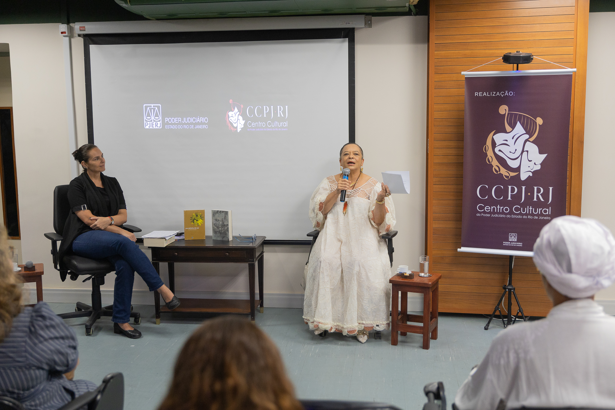 A imagem mostra um evento institucional realizado em um espaço fechado, com duas mulheres em destaque à frente de um pequeno público. Ao centro-direita, uma mulher está sentada e fala ao microfone, segurando um papel. Ela veste um traje longo, claro e rendado, com aparência elegante e tradicional. Sua expressão é séria e concentrada, indicando que está conduzindo uma fala ou leitura. À esquerda, outra mulher está sentada em uma cadeira, com postura atenta, observando a palestrante. Ela veste roupas mais informais, como blusa escura e calça jeans, sugerindo que pode ser mediadora ou participante do evento. Entre as duas, há uma mesa com alguns livros expostos. Ao fundo, uma tela de projeção exibe os logotipos do Poder Judiciário do Estado do Rio de Janeiro e do Centro Cultura. À direita, há um banner vertical com a marca do centro cultural.
