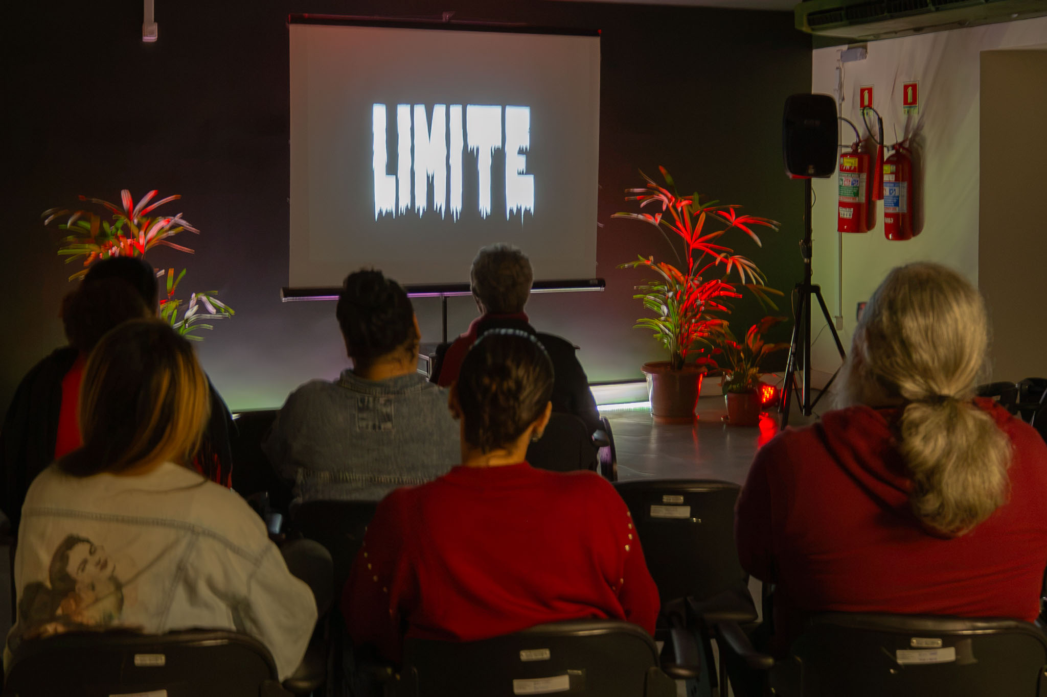 A imagem mostra uma sessão de exibição audiovisual em uma sala escura, com o público sentado de frente para uma tela de projeção. Na tela, destaca-se a palavra “LIMITE” em letras brancas estilizadas sobre fundo preto, indicando o título da obra exibida. Os espectadores estão acomodados em cadeiras organizadas em fileiras e observam atentamente a projeção. O ambiente possui iluminação reduzida, sendo complementado por luzes decorativas em tons avermelhados que iluminam plantas ornamentais posicionadas ao lado da tela. À direita da sala, há uma caixa de som sobre tripé e extintores de incêndio fixados na parede. 