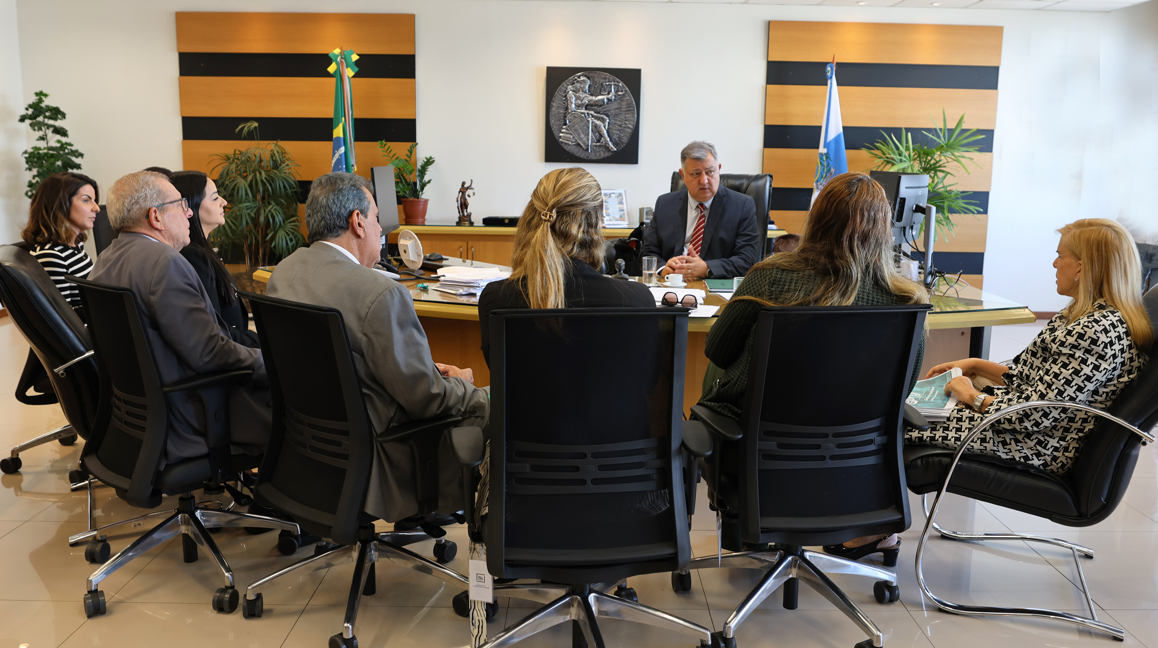 A imagem retrata uma reunião formal no gabinete corregedor-geral de Justiça, desembargador Cláudio Brandão. Ele está sentado atrás de uma mesa ampla de madeira e conduz a reunião. Ele veste terno e gravata e fala enquanto os demais participantes o escutam atentamente.À frente da mesa, há várias pessoas sentadas em cadeiras pretas, dispostas em semicírculo, a maioria também vestida de forma social. 