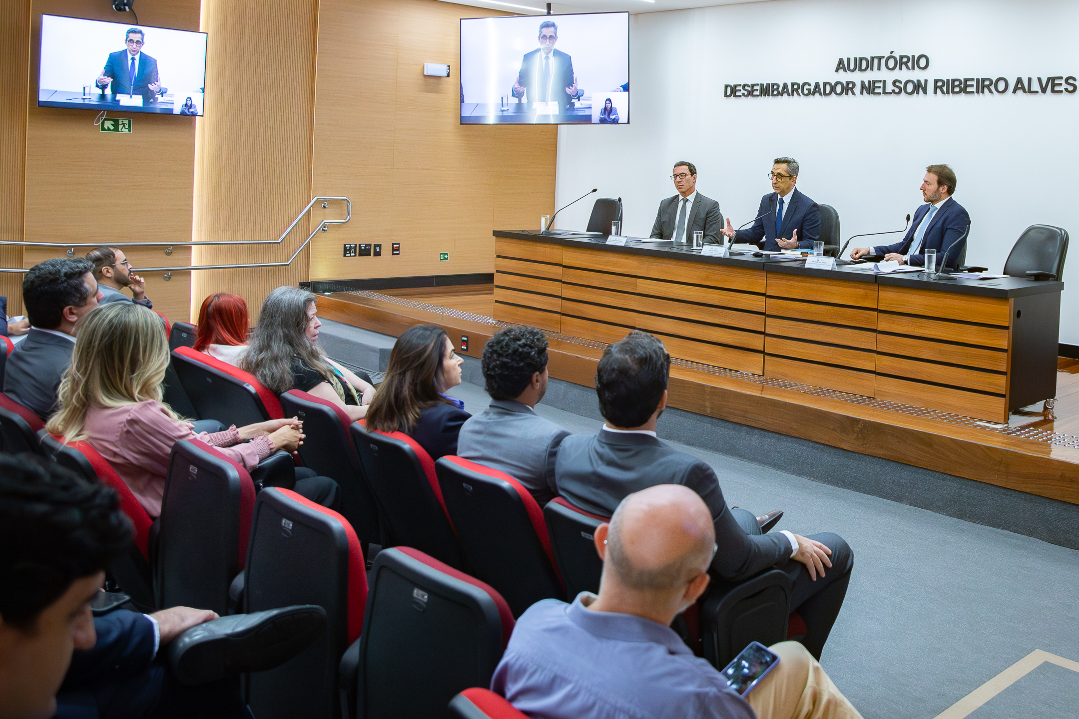 A imagem mostra um auditório durante a realização de um seminário. Em primeiro plano, o público está sentado em cadeiras estofadas, acompanhando atentamente a apresentação. À frente, três homens compõem a mesa de palestrantes, sentados atrás de uma bancada com microfones e copos d’água. Um deles, ao centro, está com as mãos levantadas, gesticulando enquanto fala, indicando que conduz a exposição no momento. Na parede ao fundo, lê-se “Auditório Desembargador Nelson Ribeiro Alves”, identificando o local. Acima da mesa, duas telas exibem a transmissão do palestrante, além de uma intérprete de Libras em destaque, indicando acessibilidade e possível transmissão ao vivo.