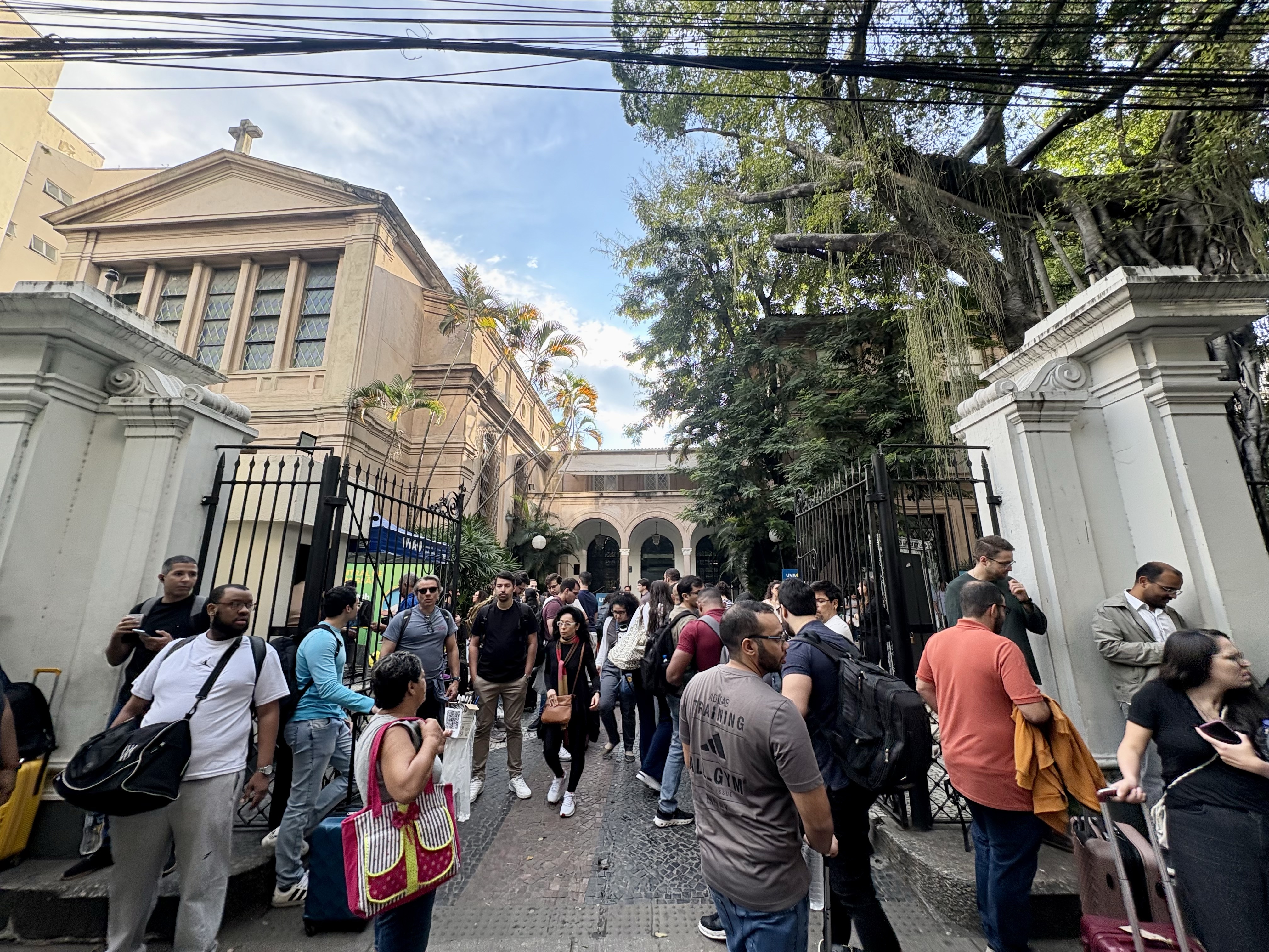 A imagem mostra a entrada de um edifício histórico, com arquitetura de inspiração clássica, cercado por um portão de ferro aberto. A fachada apresenta colunas, frontão triangular encimado por uma cruz e janelas altas com vitrais. À direita, uma grande árvore de copa extensa e raízes aéreas se destaca sobre o muro de entrada, proporcionando sombra ao local. Em primeiro plano, dezenas de pessoas circulam e permanecem reunidas na calçada e na área de acesso ao prédio. Algumas conversam em pequenos grupos, enquanto outras aguardam, caminham ou utilizam celulares. Muitas carregam mochilas, bolsas ou malas de viagem. Ao fundo, é possível ver um pátio interno com arcadas e vegetação. O céu está parcialmente nublado e há fiação aérea atravessando a parte superior da imagem, característica da paisagem urbana. 