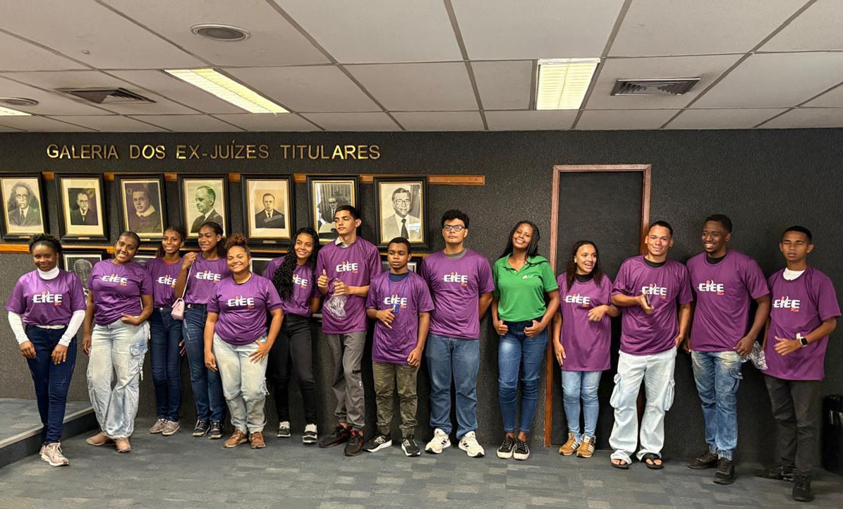 A imagem mostra um grupo de adolescentes e jovens reunidos em um ambiente interno institucional. Eles estão posicionados lado a lado, em frente a uma parede escura onde há uma galeria de retratos emoldurados e a inscrição “GALERIA DOS EX-JUÍZES TITULARES”. A maioria dos participantes veste camisetas roxas com a palavra “EJEE” estampada em branco. Entre eles, há duas mulheres usando roupas diferentes: uma veste camisa verde e jeans, e outra usa roupa escura. Alguns jovens seguram garrafas de água e fazem gestos descontraídos, enquanto sorriem para a câmera.