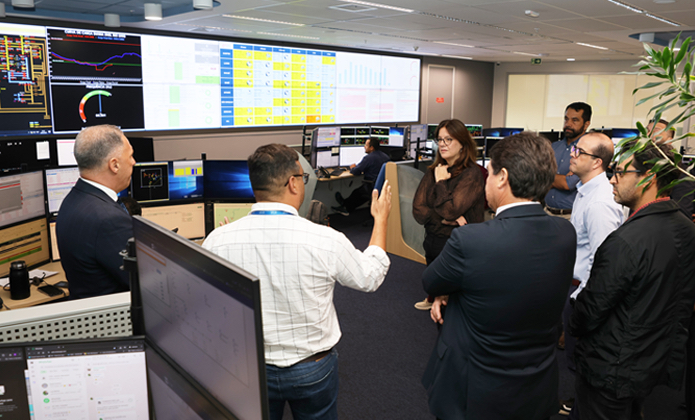 A imagem mostra um grupo de pessoas reunidas em uma sala de monitoramento. O ambiente é moderno, com diversas estações de trabalho equipadas com computadores e múltiplos monitores. Na parede ao fundo há um grande painel composto por várias telas que exibem gráficos, indicadores, tabelas e painéis de controle coloridos. No centro da cena, um homem de camisa clara, de costas para a câmera, gesticula com a mão levantada enquanto parece explicar algo ao grupo. À frente dele estão outros homens e uma mulher, todos vestidos de forma formal— alguns de terno, outros de camisa social — observando atentamente a apresentação.  Ao fundo, outras pessoas trabalham sentadas em frente aos computadores. 