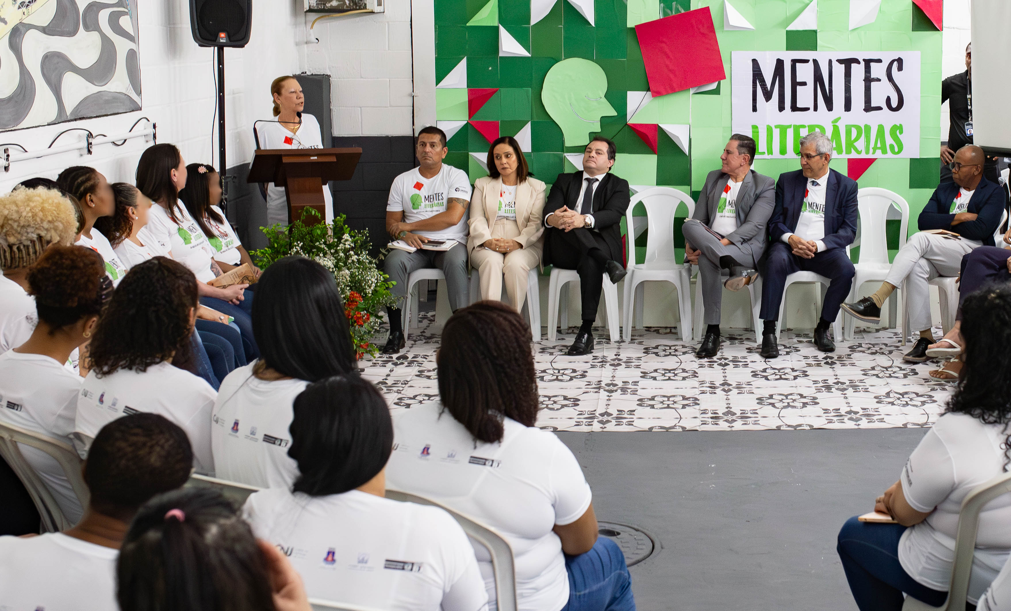 A foto apresenta as internas de costas e as autoridades sentadas de frente com a 2ª vice-presidente do TJRJ, desembargadora Maria Angélica, em pé discursando.