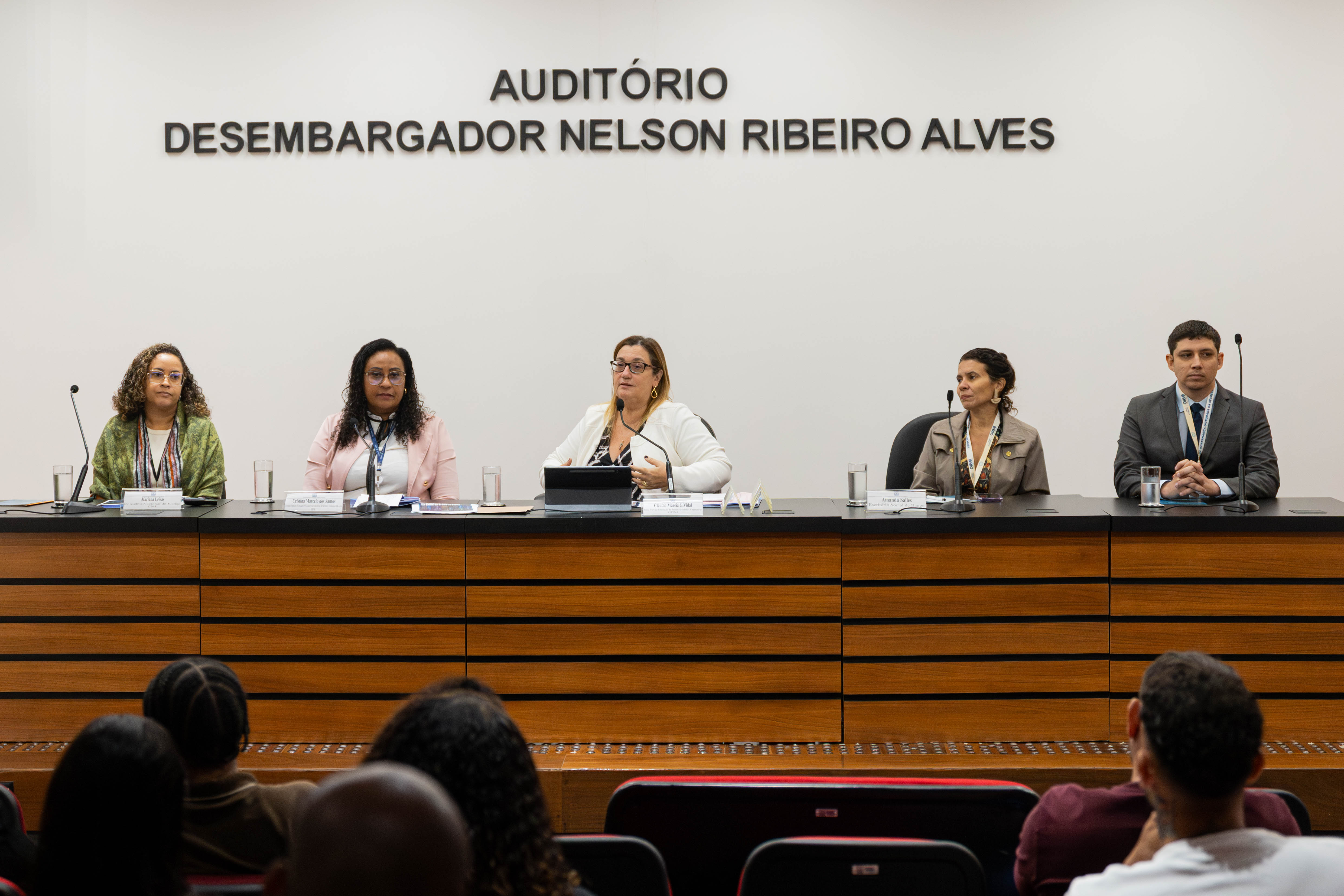 A imagem mostra um auditório durante um evento institucional. No palco, há uma mesa longa de madeira onde seis mulheres estão sentadas lado a lado, participando de uma mesa de debate. Algumas delas utilizam microfones, e há copos de água e materiais de apoio sobre a mesa. Ao fundo, na parede branca, está escrito em letras grandes: “AUDITÓRIO DESEMBARGADOR NELSON RIBEIRO ALVES”, indicando o nome do local. As participantes apresentam posturas atentas e sérias. O público aparece em primeiro plano, sentado em cadeiras de auditório, de costas para a câmera. Uma pessoa na plateia segura um celular, aparentemente registrando o momento.