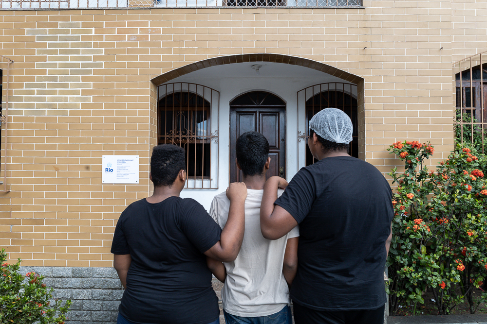 Trio de jovens posam de costas para foto. Eles estão virados para a entrada de uma casa