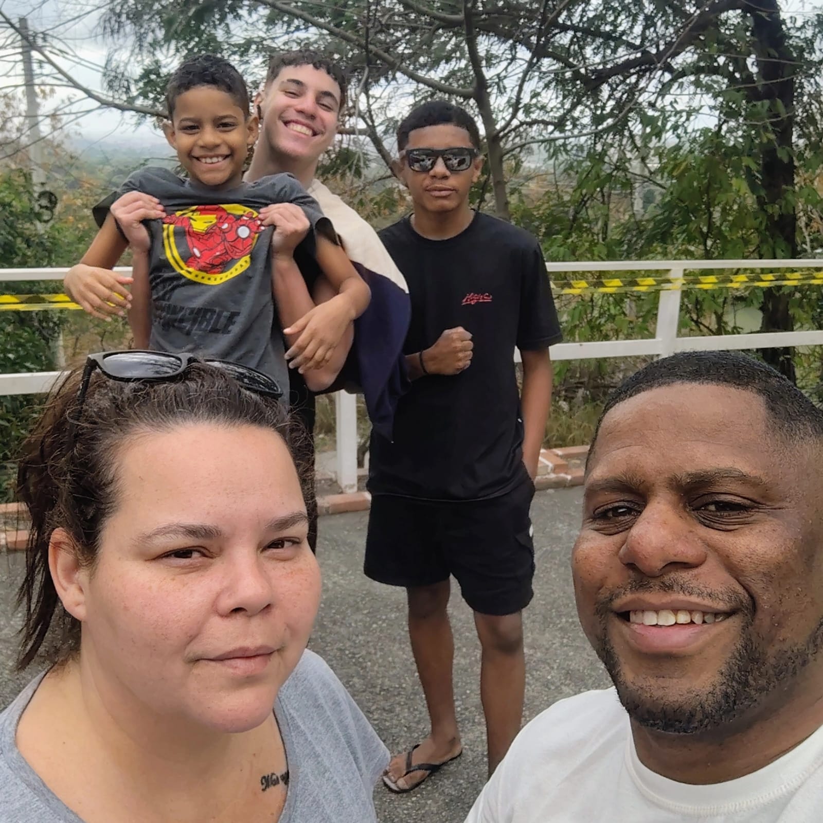 Foto mostra três meninos de diferentes idades atrás de um  homem e uma mulher. O grupo forma uma família e está em um cenário com árvores ao fundo