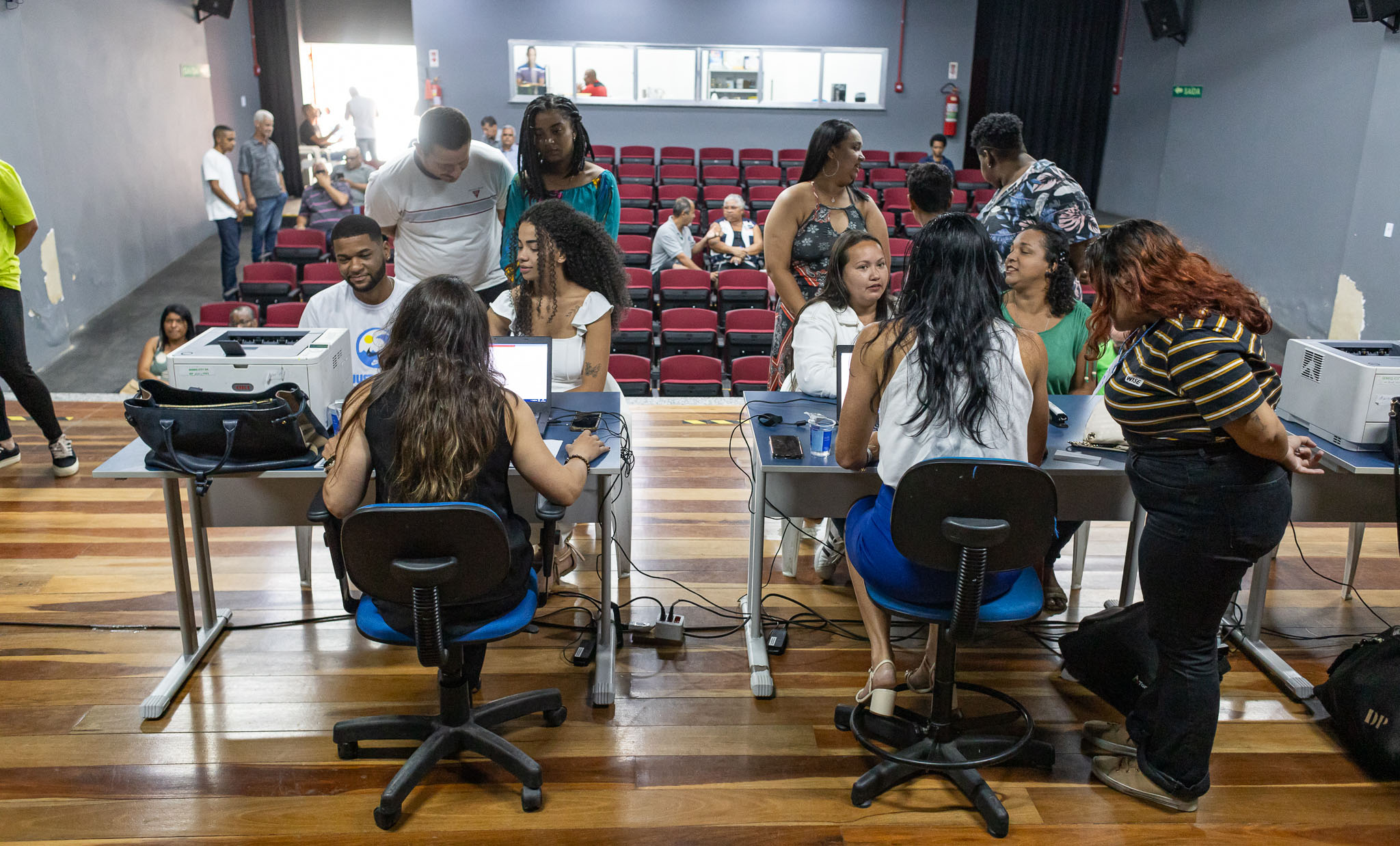 A foto mostra um grupo de pessoas no palco de um teatro usando computadores dispostos em mesas. O ambiente tem piso de madeira e paredes cinza escuro. Há várias pessoas sentadas na plateia