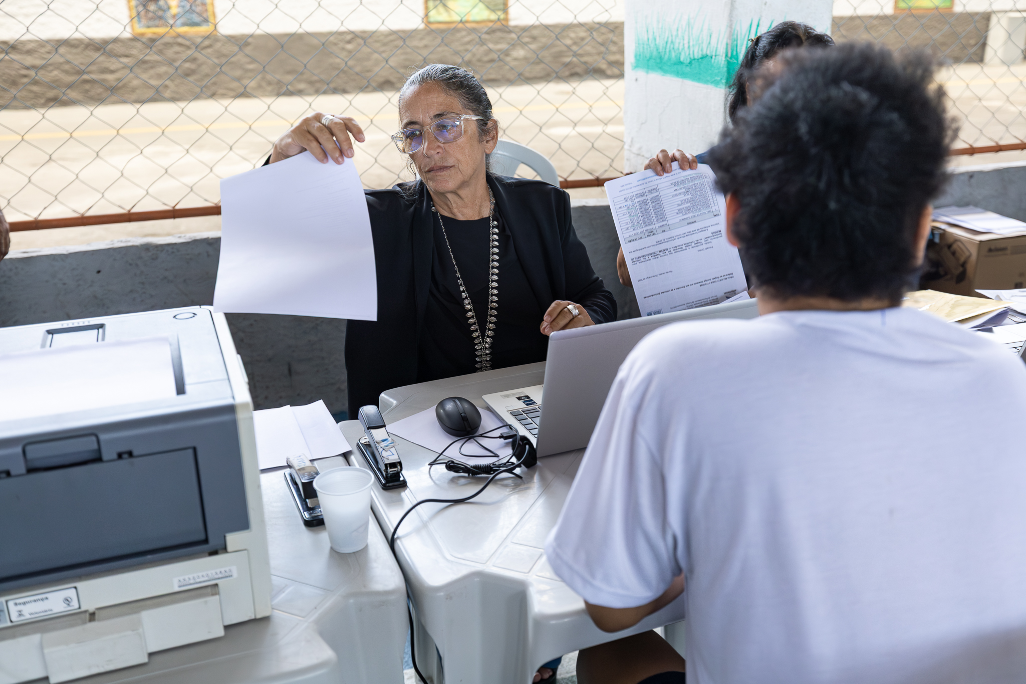 A imagem mostra um atendimento sendo realizado em um ambiente adaptado. Em primeiro plano, a desembargadora Claudia Motta sentada atrás de uma mesa entrega uma folha de papel a um homem, que está de costas para a câmera. Ela usa óculos e roupas escuras, e demonstra concentração na atividade. Sobre a mesa, há um notebook, impressora, mouse, grampeador e outros materiais de escritório. Ao lado, outra pessoa segura um documento, auxiliando no processo.