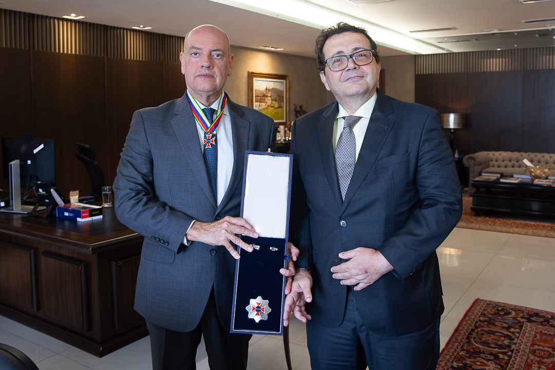Na imagem aparecem o presidente do TJRJ, desembargador Ricardo Couto de Castro (à esquerda), e o presidente do TRT-RJ, desembargador Roque Lucarelli Dattoli (à direita). O presidente do TJRJ está usando um terno cinza, camisa clara e gravata, e traz no pescoço uma medalha presa a uma fita colorida. Ele segura uma caixa aberta que exibe uma insígnia/medalha em formato de estrela. O presidente do TRT-RJ veste um terno escuro, camisa clara e gravata, e está posicionado de forma próxima, com postura formal, olhando para a câmera.