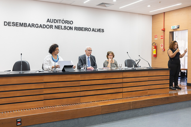 Imagem no Auditório Desembargador Nelson Ribeiro Alves. Mesa do evento formada, da esquerda para direita, pela professora Milena Afonso, desembargador Wagner Cinelli e professora Marcia Bernardes.