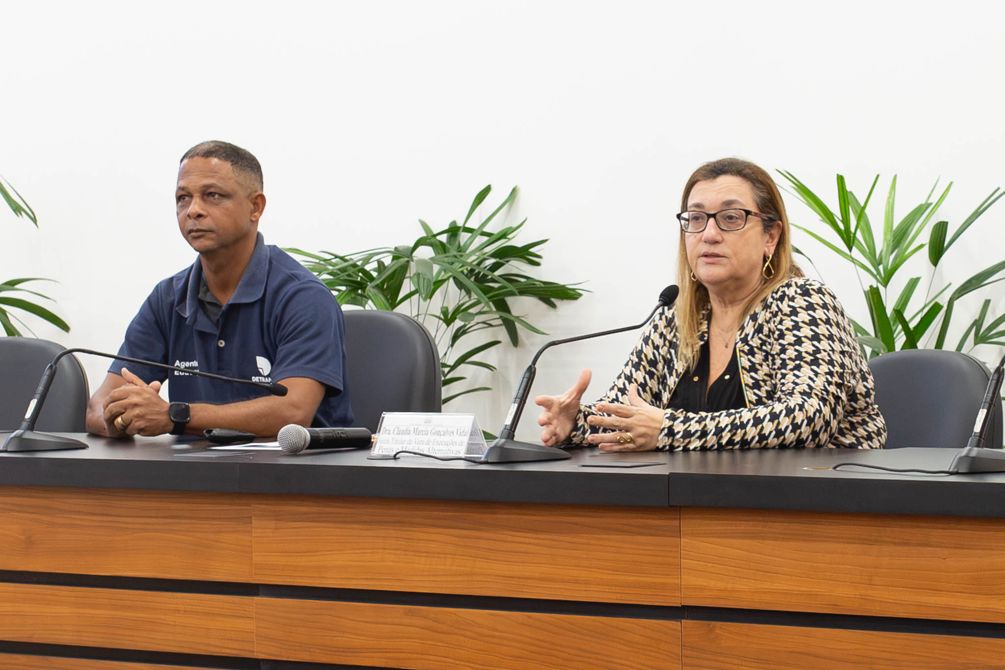 A imagem mostra dois palestrantes sentados à mesa principal de um auditório durante um evento. À esquerda, o representante do Detran Marcos dos Santos Baptista, um homem veste uma camisa polo azul-marinho, mantém as mãos apoiadas sobre a mesa e observa atentamente o ambiente. À direita, a juíza Claúdia Márcia Gonçalves Vidal, uma mulher de óculos, usando um casaco estampado sobre uma blusa preta, fala ao microfone enquanto gesticula com as mãos, demonstrando estar conduzindo a apresentação. Sobre a mesa há microfones, um microfone de mão e uma identificação da palestrante. Ao fundo, plantas ornamentais decoram o espaço diante de uma parede branca.