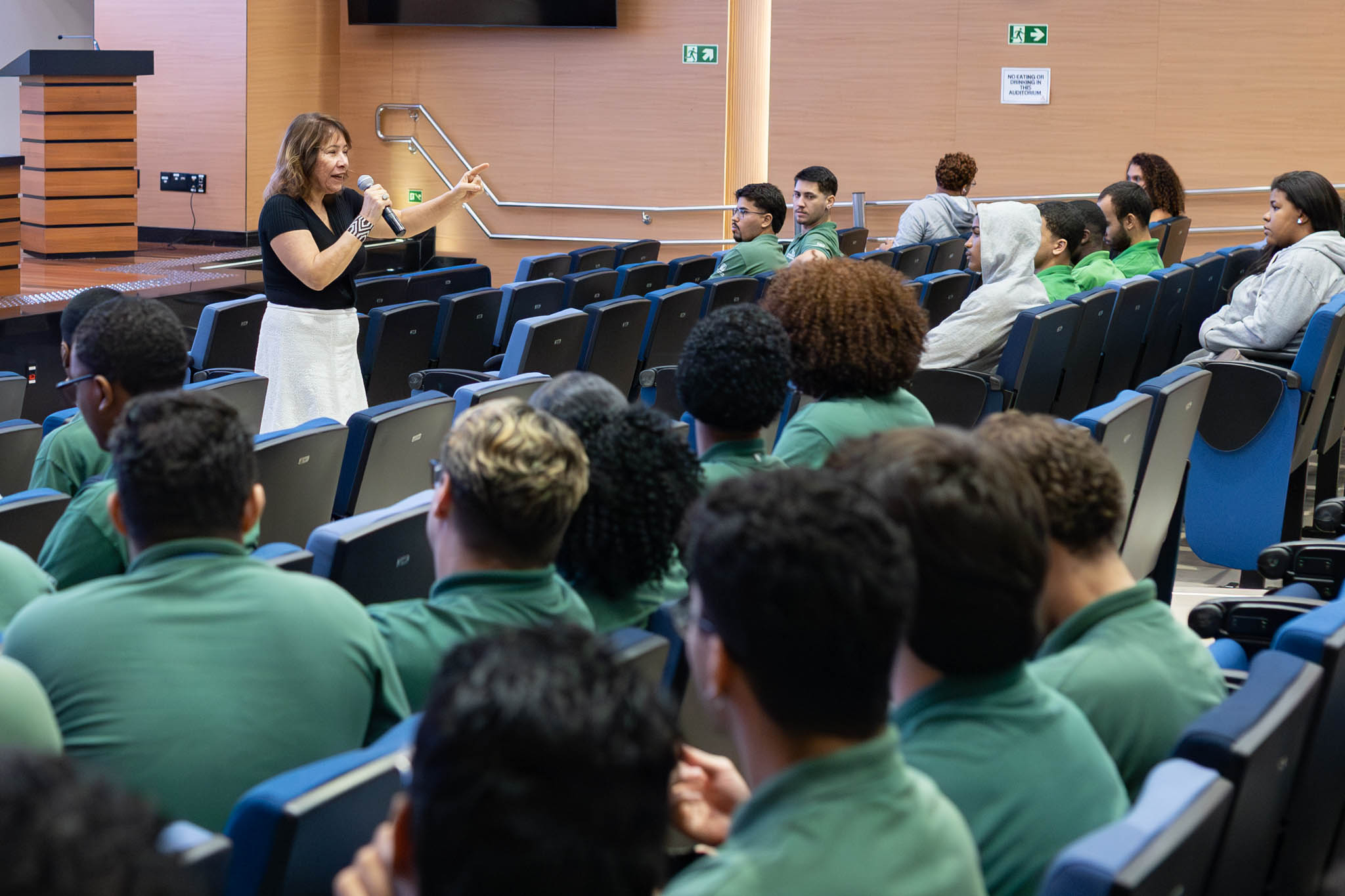 A imagem mostra uma mulher em pé, em um auditório, falando ao público com um microfone na mão. Ela veste blusa preta e saia clara e gesticula com o braço estendido, apontando para alguém na plateia, indicando interação direta com os participantes. O público é composto majoritariamente por jovens sentados em cadeiras azuis, muitos usando camisetas verdes, atentos à fala da palestrante. Alguns olham diretamente para ela, enquanto outros acompanham com expressões de interesse. O ambiente é um auditório moderno, com iluminação clara, paredes em tons neutros e estrutura organizada em fileiras.