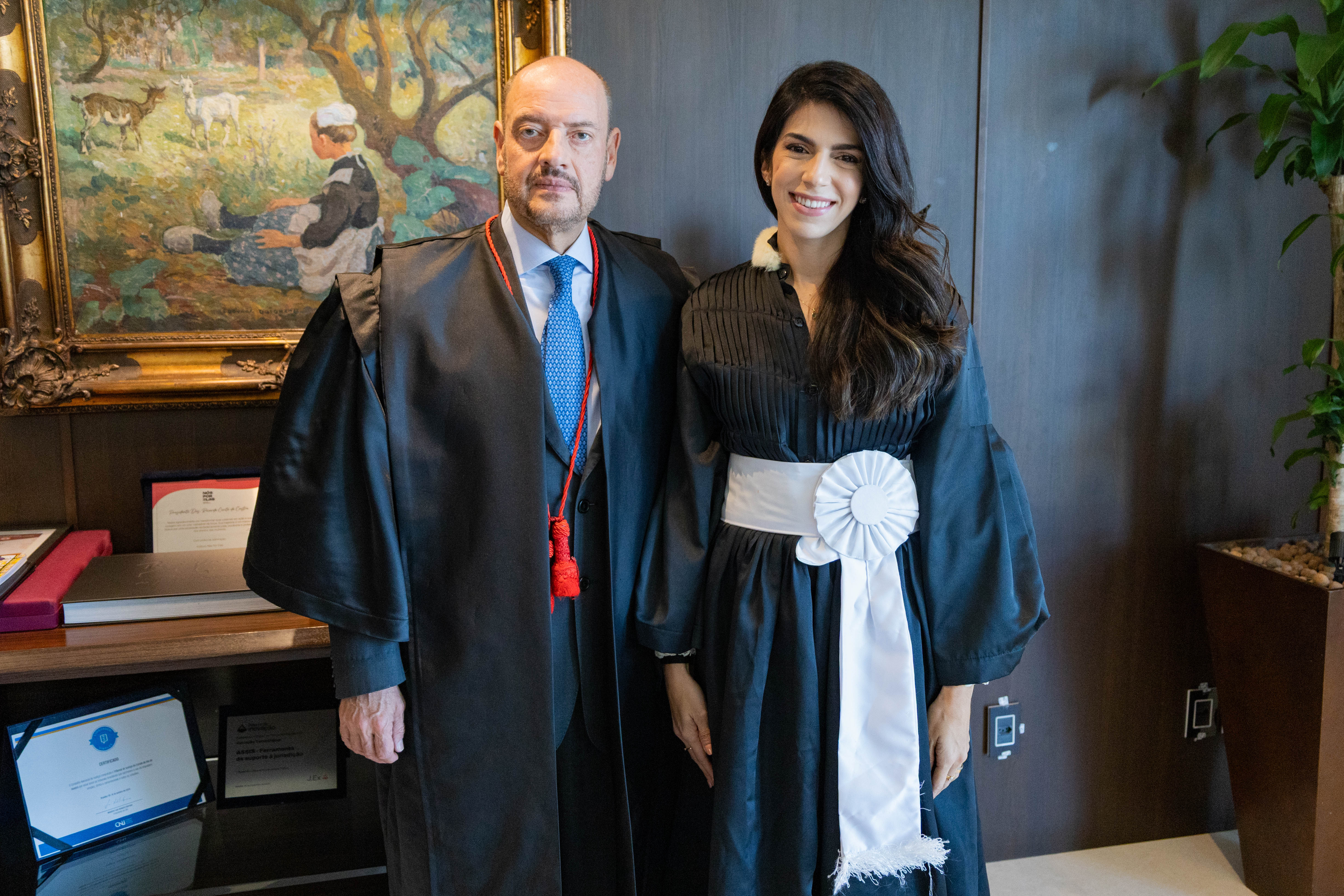 A fotografia mostra duas pessoas posando lado a lado em um ambiente institucional. À esquerda, o presidente do TJRJ, desembargador Ricardo Couto de Castro, que veste toga preta sobre terno escuro, camisa branca e gravata azul, com um cordão vermelho característico da vestimenta da magistratura. À direita, a juíza lanna Rosa Dantas Lents, uma mulher sorri para a câmera usando toga preta com jabô branco e faixa branca na cintura, também traje tradicional utilizado por magistrados em cerimônias oficiais. Ao fundo, há um painel de madeira escura. À esquerda da imagem, uma mesa exibe diplomas, placas de homenagem e outros objetos decorativos. Acima dela, um quadro emoldurado retrata uma cena campestre, com uma pessoa sentada sob árvores e animais ao redor. À direita, um vaso com uma planta de folhas verdes completa o ambiente.