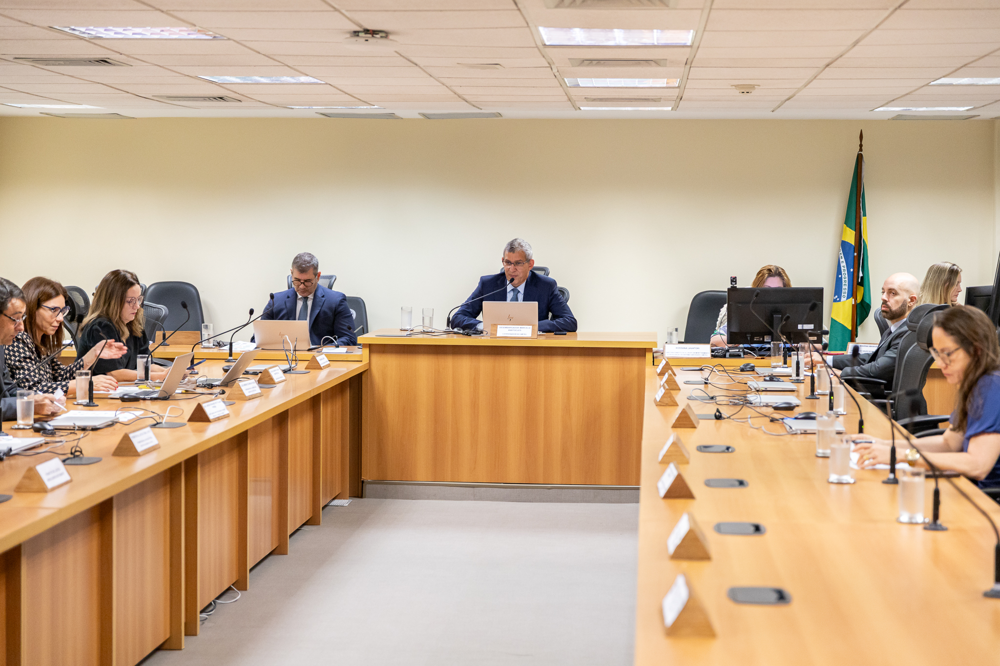 Foto de homens e mulheres reunidos em mesa em forma de U, com microfones e computadores à frente da maioria, sugerindo uma reunião em sala com pintura clara. Um homem de terno escuro está sentado na parte principal, ao centro da mesa 