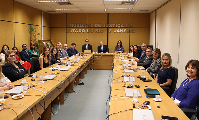 A imagem mostra uma reunião formal realizada em uma sala de conferência do Tribunal de Justiça do Estado do Rio de Janeiro (TJRJ). A sala é ampla, com paredes revestidas de madeira clara e iluminação fluorescente no teto. Ao fundo, na parede central, está o letreiro metálico com o nome do Tribunal.  Há cerca de 24 pessoas sentadas ao redor de uma grande mesa retangular de madeira clara, divididas igualmente dos dois lados. Todos estão vestidos de maneira formal — ternos, vestidos sociais, blazers — o que sugere um encontro institucional ou de trabalho. No centro, na cabeceira da mesa, estão três homens sentados, possivelmente os responsáveis por conduzir a reunião. À frente de cada participante há microfones, xícaras de café, copos com água, pratinhos com biscoitos e cadernos ou pastas. Algumas pessoas sorriem para a foto, que parece ter sido registrada de maneira oficial durante ou logo após o encontro.  O clima geral é cordial e profissional.