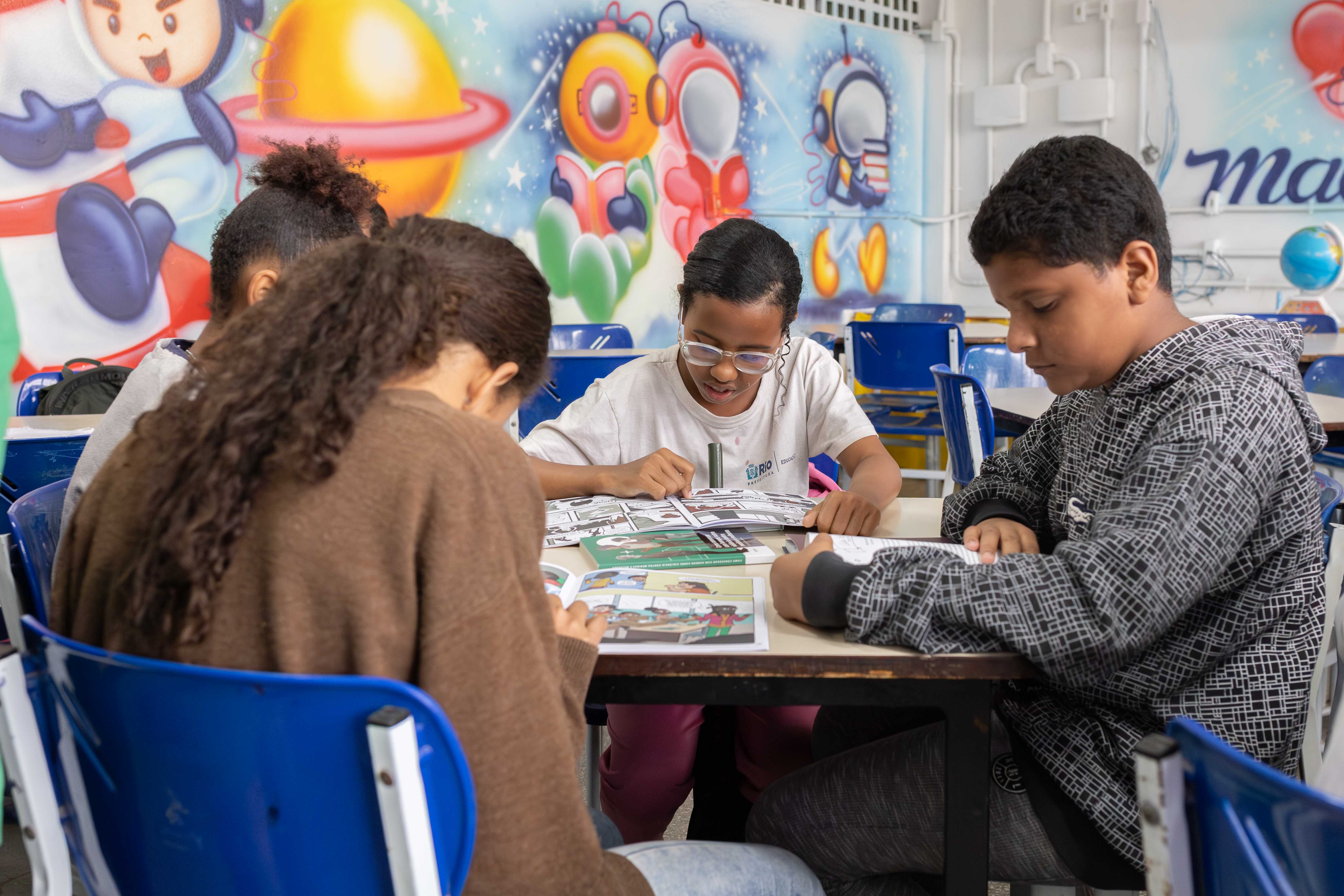 Alunos sentados em volta de uma mesa lendo livros no Espaço Maria da Penha