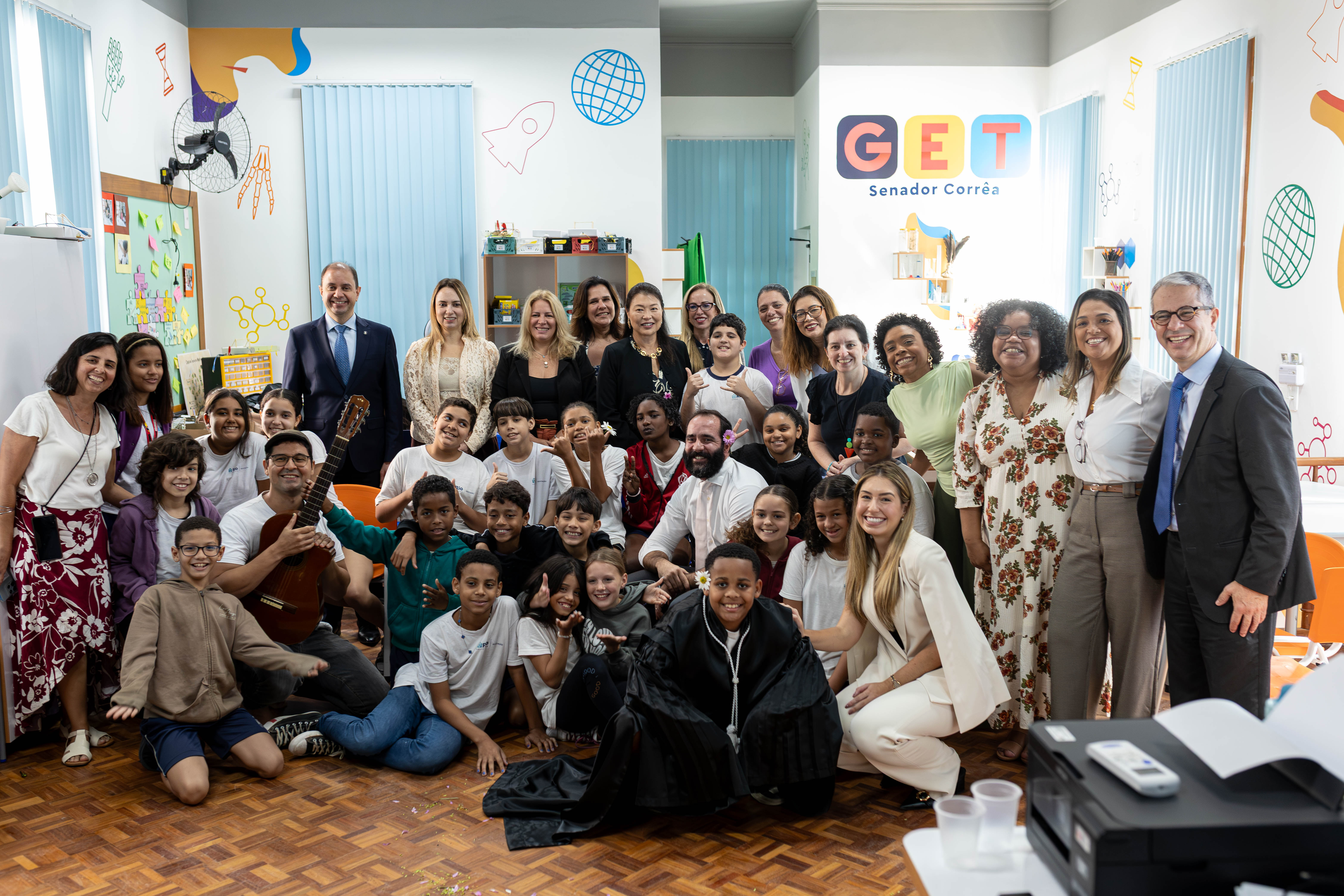 A fotografia mostra um grande grupo de adultos e crianças reunidos para uma foto em um ambiente interno colorido e voltado para atividades educacionais. Ao fundo, em uma parede branca decorada com ilustrações de foguete, globo, moléculas e outros elementos lúdicos, destaca-se a identidade visual com a inscrição "GET Senador Corrêa". As crianças ocupam a parte da frente da imagem, algumas sentadas no chão de madeira e outras ajoelhadas ou em pé. Muitas sorriem para a câmera e fazem gestos descontraídos com as mãos. Um homem sentado à esquerda segura um violão, enquanto outro, no centro do grupo, está ajoelhado entre as crianças. Um dos meninos, em primeiro plano, veste uma beca preta, destacando-se dos demais. Na fileira de trás, diversos adultos, entre eles homens de terno e mulheres com trajes sociais e casuais, posam lado a lado, todos voltados para a câmera e sorrindo. À direita da imagem, um homem de terno escuro, camisa branca e gravata azul está inclinado em direção ao grupo, também sorrindo. O ambiente é bem iluminado por grandes janelas com persianas verticais azuis. Há estantes com materiais pedagógicos, um mural com papéis coloridos e outros recursos educativos distribuídos pelo espaço, reforçando o caráter acolhedor e voltado ao aprendizado.