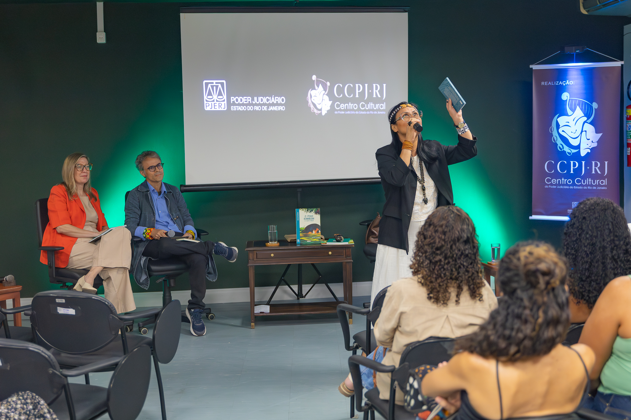 A imagem mostra um evento cultural realizado em um auditório, com uma mesa de debate posicionada à frente do público. Em destaque, uma mulher em pé fala ao microfone enquanto levanta um livro com uma das mãos, direcionando-o à plateia. Ela veste roupas claras com um blazer escuro e acessórios, transmitindo uma postura expressiva e envolvente. Ao fundo, um telão exibe os logotipos do Poder Judiciário do Estado do Rio de Janeiro (PJERJ) e do CCPJ-RJ (Centro Cultural do Poder Judiciário). A iluminação verde nas laterais cria um ambiente moderno e institucional. À direita, há um banner vertical com a identidade visual do CCPJ-RJ. Sentados ao lado, dois mediadores acompanham a apresentação: uma mulher com blazer vermelho e calça clara, segurando um caderno, e um homem com roupa casual — camisa, jaqueta e tênis — também atento. Entre eles, uma pequena mesa de madeira exibe um livro em destaque, além de um copo de água. A plateia, composta majoritariamente por mulheres, está sentada em cadeiras voltadas para o palco, observando atentamente a fala. 
