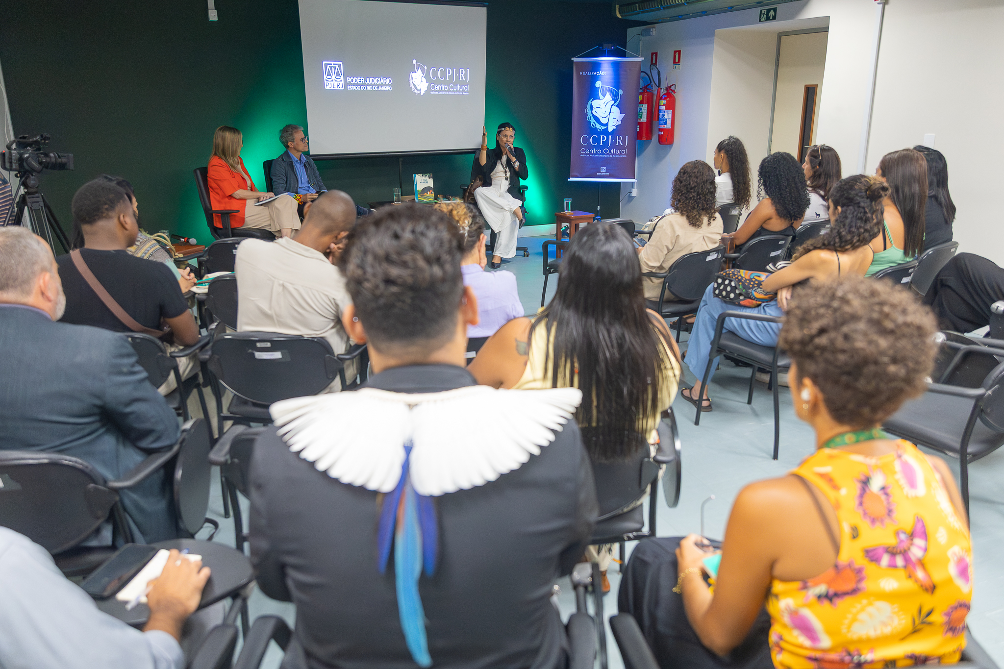 A imagem mostra um evento realizado em um auditório de pequeno porte, com o público sentado em cadeiras voltadas para um palco simples. No centro, há uma mesa com três participantes: uma mulher vestindo roupa clara, que está falando ao microfone e gesticulando com a mão levantada, e dois mediadores ao lado, um homem e uma mulher, sentados e atentos. Ao fundo, um telão exibe a identidade visual do evento, com os logotipos do Poder Judiciário do Estado do Rio de Janeiro e do CCPJ-RJ (Centro Cultural do Poder Judiciário). Ao lado direito do palco, há um banner vertical com a mesma identidade visual. A plateia é diversa, composta por homens e mulheres, muitos deles atentos à fala, alguns fazendo anotações. 