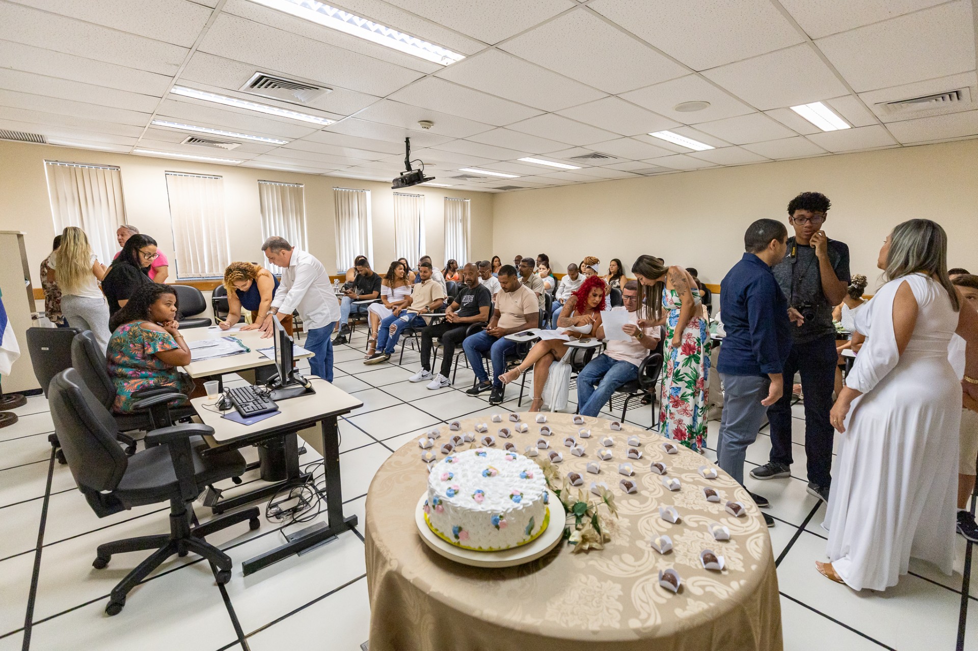 A imagem mostra uma sala ampla e bem iluminada, com muitas pessoas reunidas. No primeiro plano, há uma mesa redonda coberta por uma toalha bege, sobre a qual está um bolo decorado com glacê branco e detalhes coloridos. Pequenos doces embalados estão organizados ao redor do bolo.  No fundo, várias pessoas estão sentadas em cadeiras enfileiradas, segurando papéis e aparentando estar em um evento formal. Algumas estão conversando, enquanto outras parecem estar preenchendo ou lendo documentos. Há uma diversidade de roupas, incluindo trajes mais casuais e vestidos floridos.  À esquerda da imagem, uma mulher está sentada atrás de uma mesa com um computador, parecendo trabalhar ou organizar algo. Ao lado dela, outra mulher e um homem de camisa branca estão assinando ou verificando papéis.  Na parte direita, algumas pessoas estão de pé e conversando. Uma mulher vestindo branco longo parece estar interagindo com um homem de camisa azul e outro jovem de roupa escura.  O ambiente tem um ar institucional, com paredes claras e janelas cobertas por persianas. O teto possui iluminação fluorescente e há um projetor pendurado. A cena sugere que o evento pode estar relacionado a um casamento comunitário ou algum tipo de cerimônia formal, possivelmente no fórum de Madureira, como indicado no nome do arquivo.