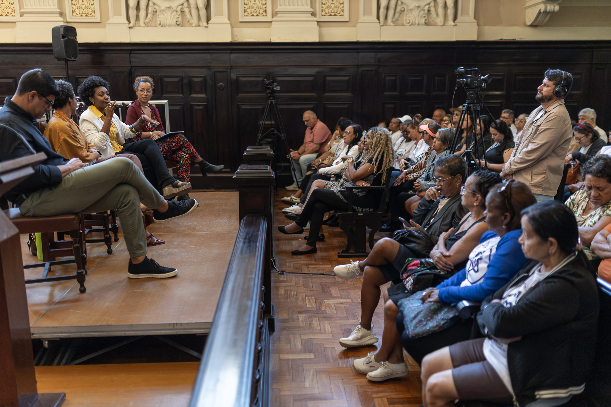 Em um salão histórico com piso de madeira, painéis escuros e detalhes arquitetônicos ornamentados nas paredes, uma mesa de debate reúne cinco participantes sentados sobre um palco. Ao centro, uma das palestrantes, vestindo blazer branco e blusa amarela, fala ao microfone enquanto gesticula com uma das mãos. Os demais integrantes da mesa acompanham a exposição, alguns observando a palestrante e outros consultando anotações. À frente do palco, o público ocupa fileiras de bancos e cadeiras, acompanhando atentamente a discussão. Entre os presentes há pessoas de diferentes idades, algumas utilizando celulares ou fazendo anotações. À direita da imagem, um cinegrafista registra o evento com uma câmera posicionada sobre um tripé. Ao fundo, outra câmera também está instalada para gravar a atividade.