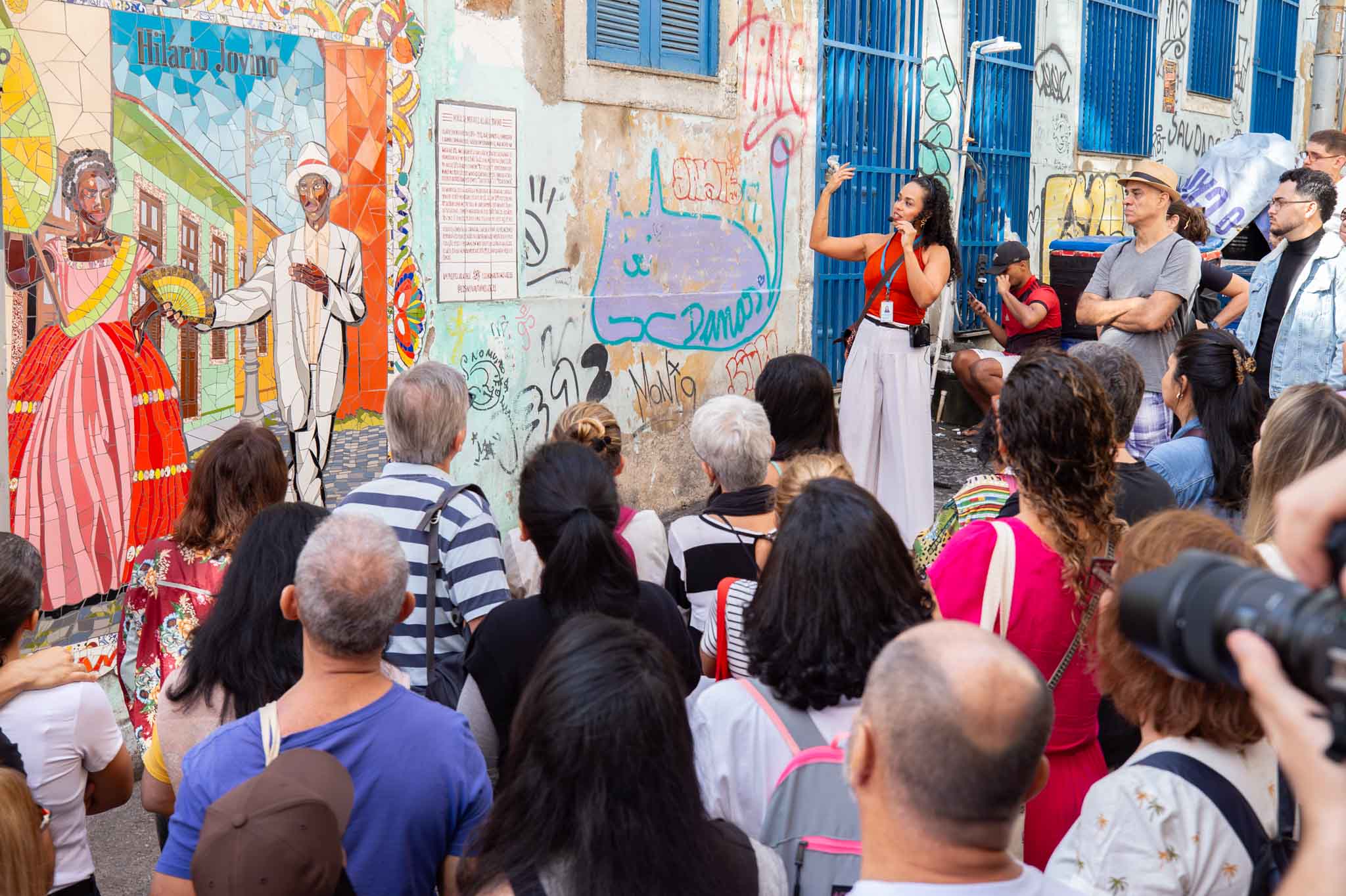 A imagem mostra um grupo de pessoas reunidas em uma atividade cultural e histórica ao ar livre, em uma rua da região da Pequena África, no Centro do Rio de Janeiro. Em primeiro plano, o público aparece de costas para a câmera, observando uma guia que conduz a atividade. A mulher, vestida com blusa vermelha e saia clara, gesticula enquanto fala para os participantes. À esquerda da imagem, há um grande mural colorido em mosaico representando personagens históricos negros em trajes tradicionais. Acima da obra aparece a inscrição “Hilário Jovino”. A parede ao redor do mural possui grafites e intervenções urbanas. Alguns participantes acompanham atentamente a explicação, enquanto outros observam o painel artístico. 