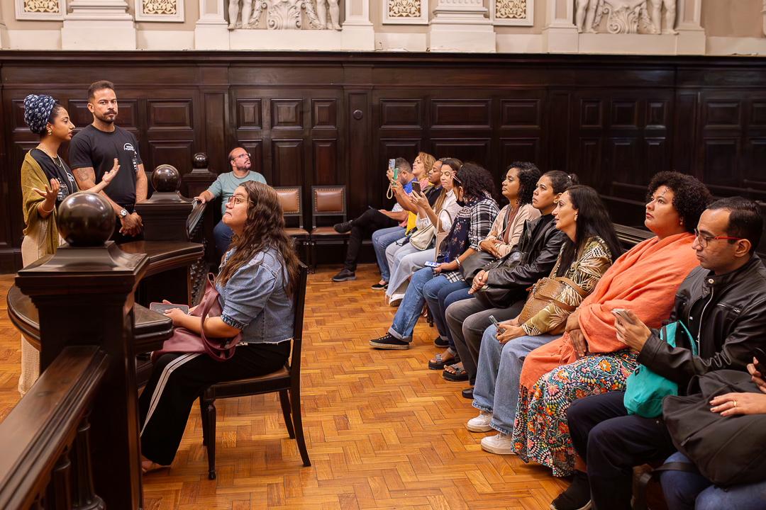 A imagem retrata um grupo de pessoas participando de uma visita guiada no salão histórico do Tribunal do Júri. O espaço é marcado por paredes de madeira escura, detalhes arquitetônicos clássicos e piso de madeira, criando uma atmosfera solene e institucional. À esquerda, duas pessoas conduzem a explicação: uma mulher gesticula enquanto fala ao grupo, e um homem permanece ao seu lado, atento. Ambos estão de pé, próximos a uma estrutura que remete à tribuna ou bancada original do espaço. À direita, os visitantes estão sentados em fileiras, observando atentamente. Alguns seguram celulares, possivelmente registrando o momento, enquanto outros acompanham a explicação com expressões de interesse. 