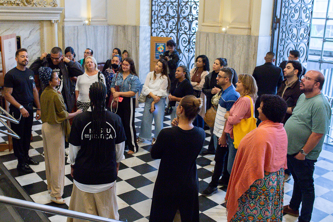 A imagem mostra um grupo de pessoas reunido em uma visita guiada dentro do Museu da Justiça. O ambiente tem piso em padrão xadrez preto e branco, paredes claras com acabamento em mármore e portas altas com grades ornamentais de ferro, que reforçam o caráter histórico do local. No centro da cena, uma mulher parece conduzir a explicação, falando para o grupo que se organiza em semicírculo, atento às orientações. As pessoas estão vestidas de forma casual e variada, algumas segurando bolsas ou objetos pessoais. 