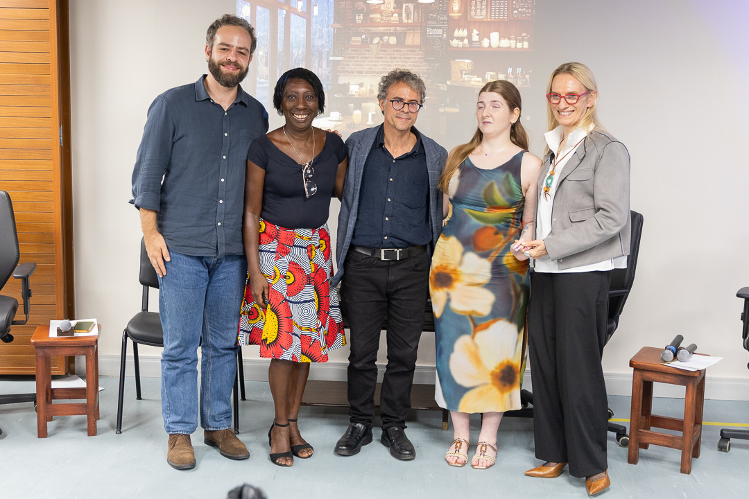 A imagem mostra cinco pessoas em pé, lado a lado, posando para uma foto em um ambiente interno. O grupo está disposto de forma alinhada e descontraída, todos voltados para a câmera. As roupas variam entre casual e social, com destaque para cores neutras combinadas a peças mais expressivas, como vestidos com estampas florais e uma saia colorida.