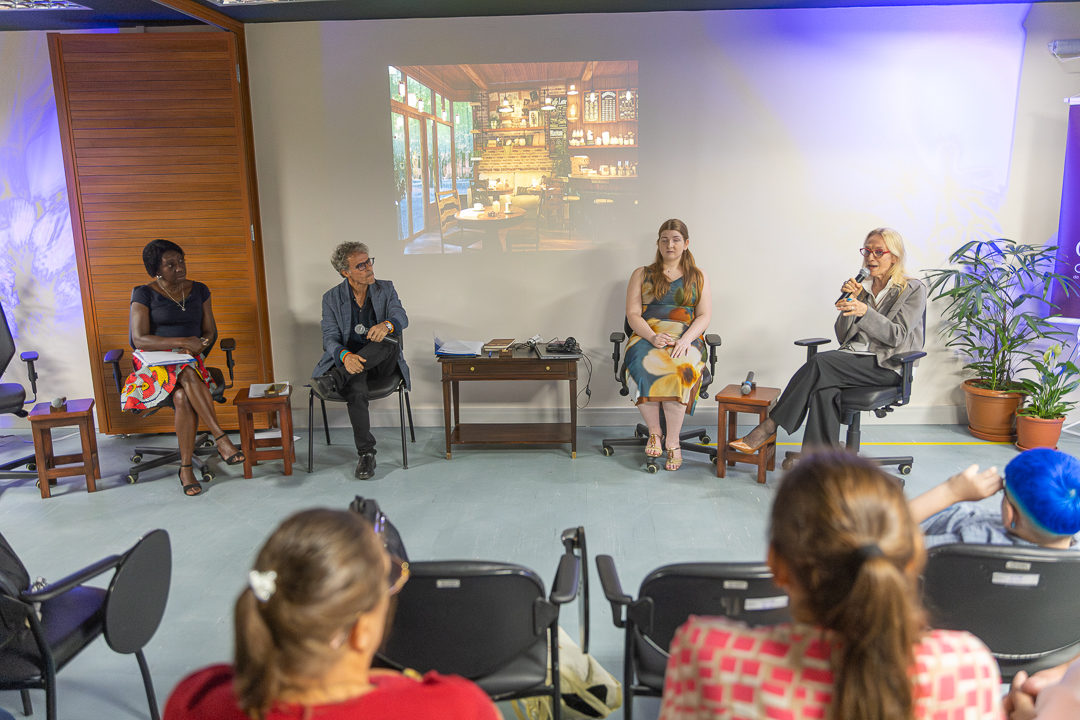 A imagem mostra um evento cultural, com formato de mesa de debate, realizado em uma sala de apresentações. Quatro pessoas estão sentadas em cadeiras, dispostas lado a lado, voltadas para o público. À direita, a desembargado Cristina Tereza Gaulia segura um microfone e fala, aparentando conduzir a discussão. Os demais integrantes da mesa a escutam com atenção, em postura tranquila e reflexiva. As pessoas vestem roupas sociais ou casuais.