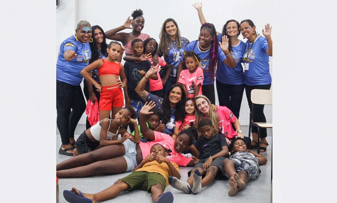 Sete recreadoras de camisa azul e calça preta posam para foto com grupo de crianças sentadas e deitadas no chão, vestindo roupas coloridas, em uma sala de paredes claras