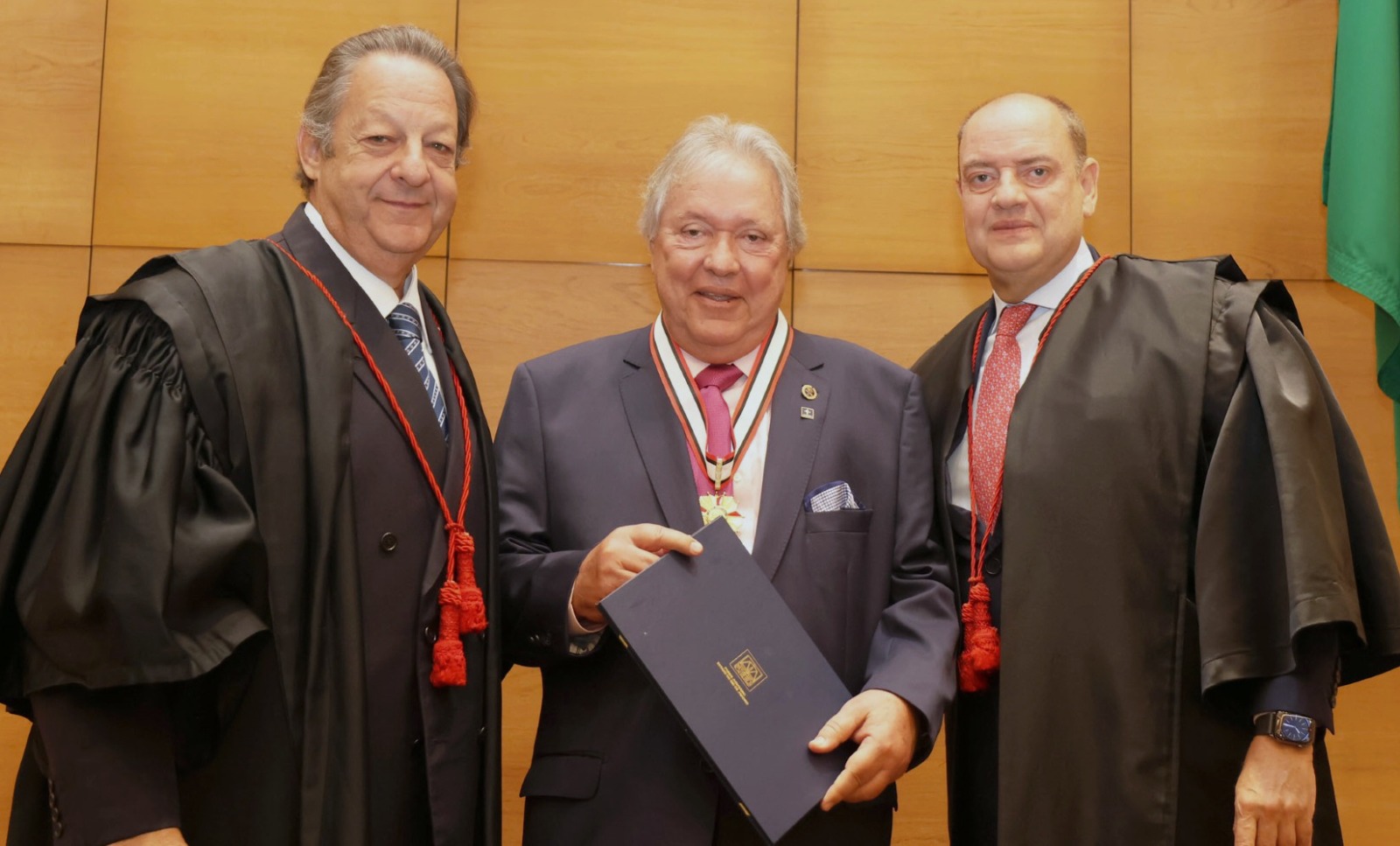 Foto mostra três homens: à esquerda está o mais alto e vestindo toga da magistratura, ao centro, o mais baixo, de cabelos brancos, de terno e com comenda pendurada no pescoço como colar e segurando diploma, e à direta, também vestindo toga, o terceiro homem, que usa gravata vermelha. 