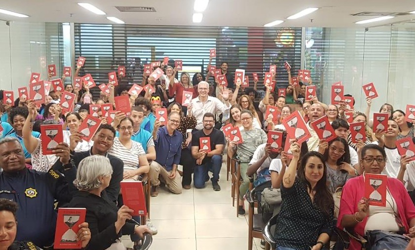 Em auditório de paredes e piso claros, dezenas de pessoas estão sentadas seguram, com braço ao alto, exemplares de livro de capa vermelha. Ao fundo, de camisa branca, o desembargador Wagner Cinelli está entre as pessoas