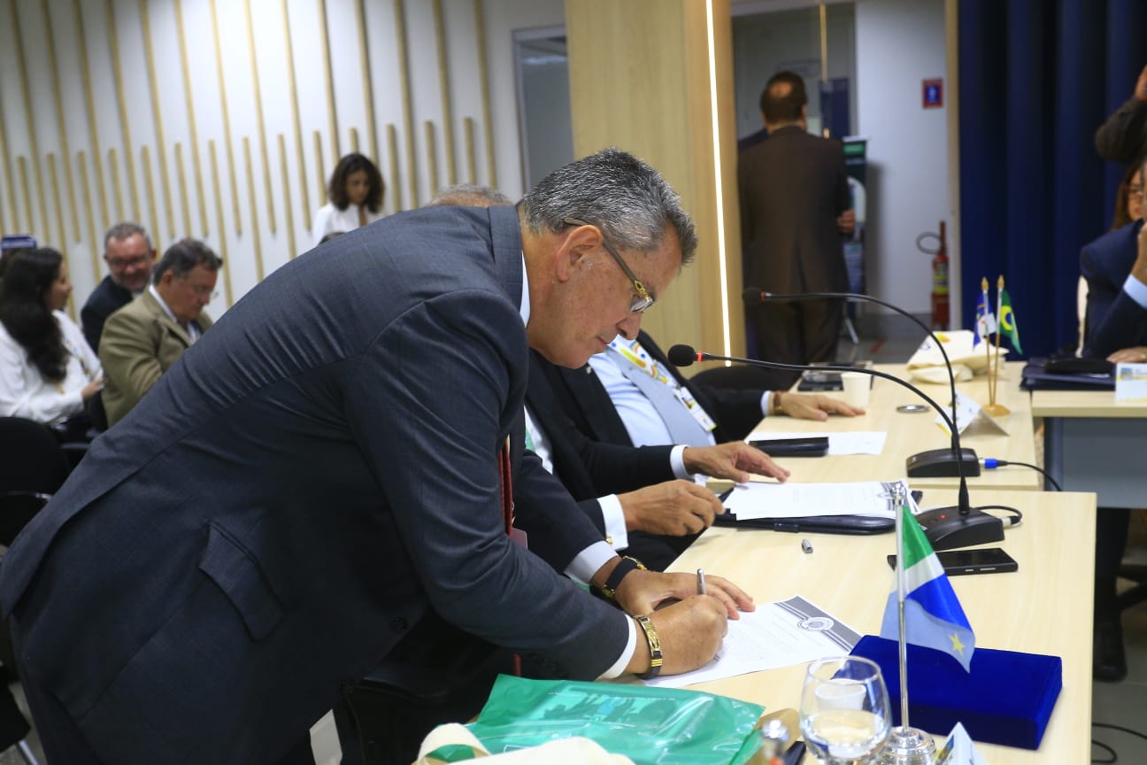 A imagem mostra o presidente do Tribunal Regional Eleitoral do Rio de Janeiro (TRE-RJ), desembargador Claudio de Mello Tavares, assinando documento durante cerimônia. O magistrado usa terno escuro, camisa branca, gravata em tom salmão e óculos, inclina-se sobre a mesa para assinar um documento com uma caneta. Ele utiliza também um crachá preso a um cordão.