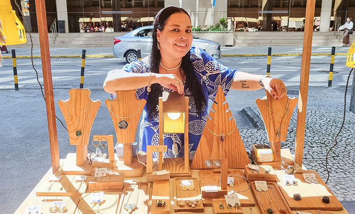 Mulher de roupa azul mostra as bijuterias expostas em uma das barracas da feira de artesanato