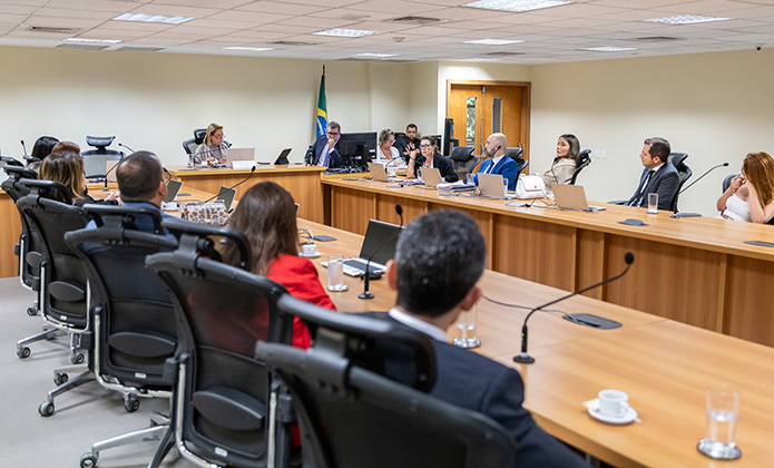 A imagem mostra um grupo de pessoas sentadas ao redor de uma mesa de madeira em uma sala de reunião 