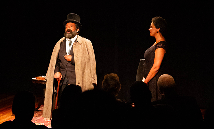 A imagem mostra uma cena de teatro encenada em um palco com iluminação dramática e fundo totalmente escuro, que foca a atenção nos dois atores em destaque. À esquerda, um homem com figurino de época — incluindo cartola, capa e bengala — interpreta seu papel com expressividade, sugerindo um personagem antigo ou histórico. À direita, uma mulher com um elegante vestido preto observa-o atentamente, parecendo compor uma cena de diálogo intenso ou solene.  Na frente da imagem, é possível ver a silhueta do público assistindo à apresentação, reforçando o ambiente cênico e a atmosfera de espetáculo.