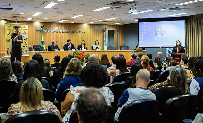 A imagem mostra um auditório lotado durante um evento formal. Na parte frontal do espaço, há uma mesa com cinco pessoas sentadas, integrantes de um painel de discussão. Atrás deles está uma parede de madeira com a inscrição “AUDITÓRIO DESEMBARGADOR JOSÉ NAVEGA CRETTON” e bandeiras — uma delas do Brasil. À direita da mesa, uma mulher está em pé em um púlpito, ao lado de um telão com a projeção da tela inicial de um computador com o logotipo do PJERJ (Poder Judiciário do Estado do Rio de Janeiro).  À esquerda do painel, há um intérprete de Libras (Língua Brasileira de Sinais). Atrás dele, um banner com o título “Acessibilidade: um direito de todos” exibe ícones relacionados à inclusão.  O público é diverso e está atento ao que é apresentado,