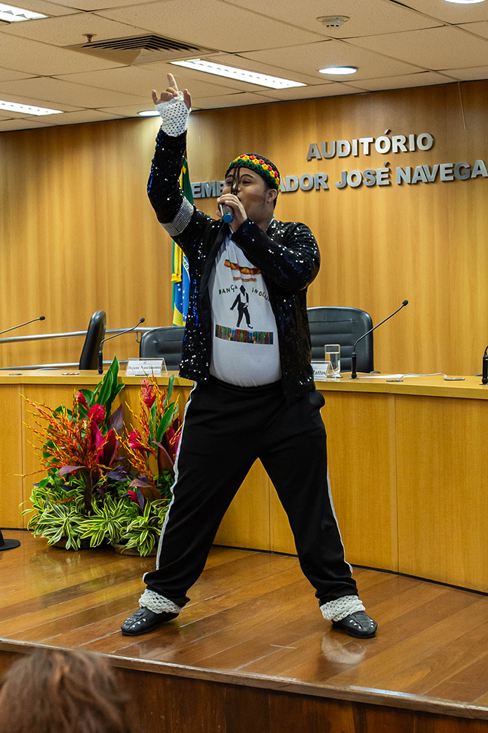 A imagem mostra um momento artístico durante o evento realizado no Auditório Desembargador José Navega Cretton. Um artista está se apresentando no palco, em frente à mesa de debates. Ele segura um microfone com a mão direita enquanto ergue a mão esquerda, fazendo um gesto expressivo, indicando entusiasmo e presença cênica.
