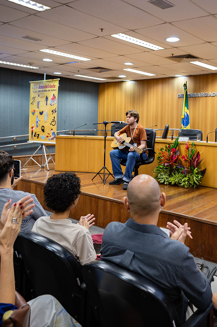A imagem mostra uma apresentação musical ocorrendo no Auditório Desembargador José Navega Cretton. Um jovem, sentado em uma cadeira sobre o palco, toca violão enquanto canta ao microfone. Ele veste uma camiseta marrom e jeans, com um crachá no pescoço.