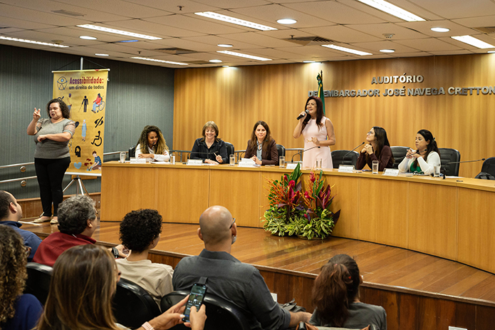 A imagem mostra um evento formal realizado em um auditório chamado “Auditório Desembargador José Navega Cretton”. Há sete mulheres sentadas ou em pé atrás de uma longa mesa em um palco de madeira. Uma das mulheres, em pé ao centro, está falando ao microfone. À esquerda da imagem, uma intérprete de Libras (Língua Brasileira de Sinais) está traduzindo para o público. Atrás dela, há um banner com o texto “Acessibilidade: um direito de todos”