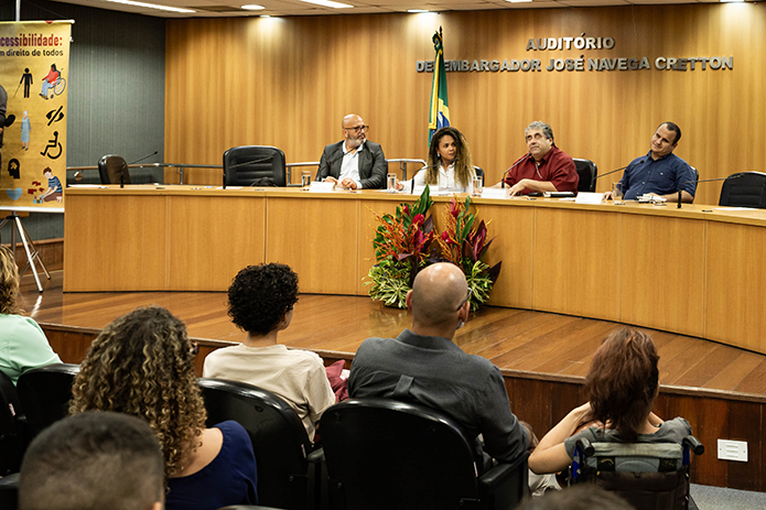 A imagem mostra uma nova composição da mesa em um auditório, o mesmo identificado como “Auditório Desembargador José Navega Cretton”. Quatro pessoas estão sentadas à mesa: duas mulheres e dois homens. Uma das mulheres, com cabelos cacheados, parece estar se preparando para falar ou ouvindo atentamente. Um dos homens, de camisa vermelha, está falando ao microfone. O ambiente é formal, com copos de água sobre a mesa e placas de identificação para cada participante.