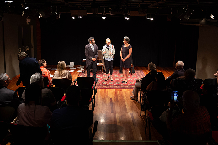 A imagem mostra um momento pós-apresentação em um teatro de pequeno porte, com três pessoas posicionadas ao centro do palco. A iluminação é focada nelas, destacando a cena. O homem à esquerda está o ator vestido com um traje formal escuro, semelhante ao figurino de época visto na imagem anterior. À direita, a atriz, vestida de preto. Ao centro, está a desembargadora Cristina Gaulia com blusa clara, calça escura e lenço no pescoço