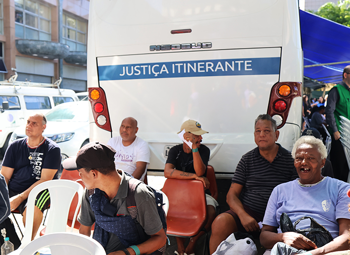 A imagem mostra seis homens sentados em cadeiras atrás do ônibus da Justiça Itinerante enquanto aguardam atendimento 