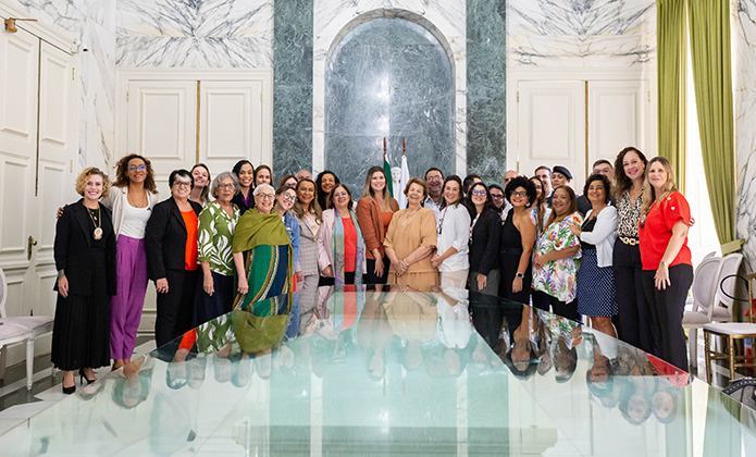 A imagem mostra um grupo diverso de aproximadamente 30 pessoas posando para uma foto coletiva em um ambiente elegante, com paredes de mármore e cortinas verdes. O grupo está disposto em semicírculo ao redor de uma grande mesa de vidro, refletindo parcialmente os participantes. A maioria das pessoas são mulheres, de diferentes idades e estilos, algumas usando roupas formais e outras mais casuais.