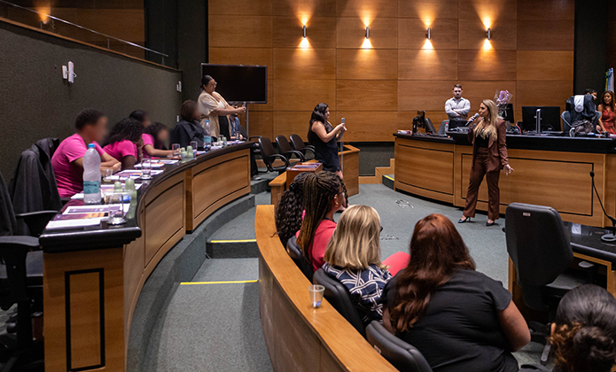 A imagem mostra o plenário, onde ocorre uma apresentação. A juíza Tula Corrêa de Mello  vestida com um terno marrom.  Na plateia, várias pessoas estão sentadas em cadeiras organizadas em semicírculo, prestando atenção. Algumas estão vestindo roupas casuais, enquanto outras usam camisetas rosa.  A iluminação do ambiente é quente, com paredes revestidas de madeira e luzes direcionadas para cima, reforçando o caráter institucional do local.