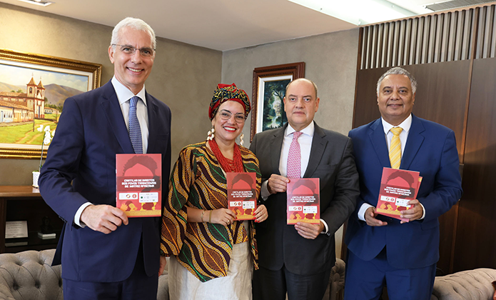 A imagem mostra quatro pessoas posando para uma foto em um ambiente formal, segurando exemplares da Cartilha dos Direitos dos Povos Tradicionais de Matriz AfricanaOs indivíduos estão bem vestidos, três homens - os desembargadores Wagner Cinelli e Ricardo Couto e o advogado  Humberto Adami - e uma mulher - Arethuza Dória - com roupas coloridas e um acessório de cabeça. 