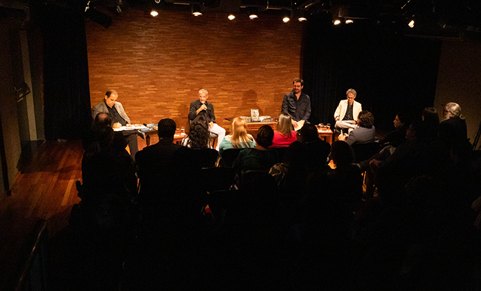 A imagem mostra quatro homens sentados em cadeiras no palco da Sala Multiuso e a plateia lotada